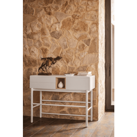 La Consola Corvo 1P2C se apoya contra una pared de piedra con su diseño elegante y funcional. Una escultura y libros apilados adornan la parte superior, y un pequeño jarrón adorna ingeniosamente el estante central abierto.