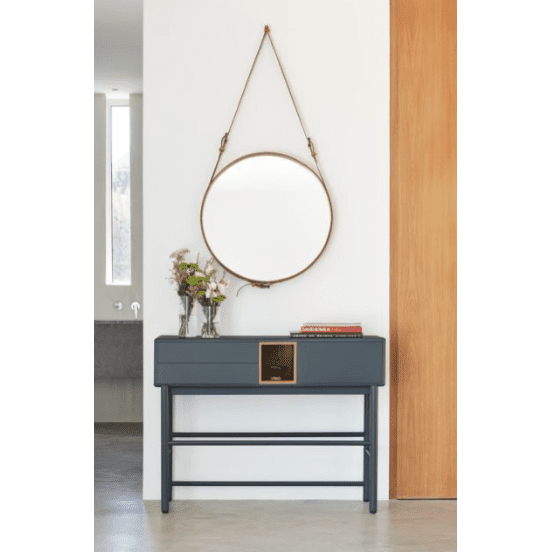 Un espejo redondo con correa de cuero cuelga sobre la Consola Corvo 1P2C, ofreciendo un diseño elegante y funcional. Un florero sobre la mesa complementa la pared blanca con detalles en madera, creando un ambiente acogedor.