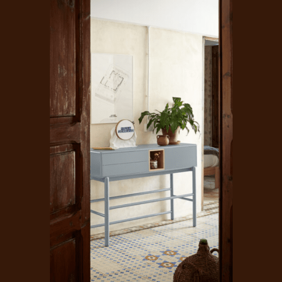 En una pequeña habitación, una mesa azul y una maceta con plantas acompañan la elegante solución de almacenaje Consola Corvo 1P2C. Un suelo de baldosas estampadas realza el ambiente, mientras que una puerta de madera enmarca la escena en primer plano.