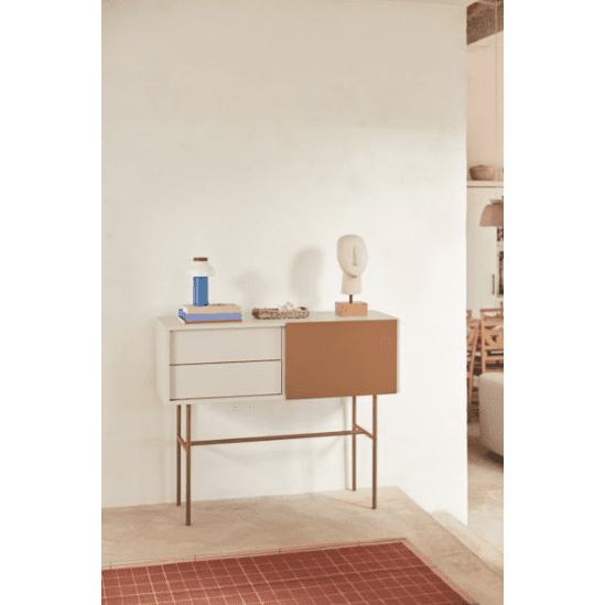 La Consola Nube 1P2C, con su diseño minimalista y paneles blancos y marrones, se alza sobre una pared clara. Está adornada con un libro azul, una canasta y una escultura abstracta blanca, mientras que un tapete rojo acentúa su estilo contemporáneo.