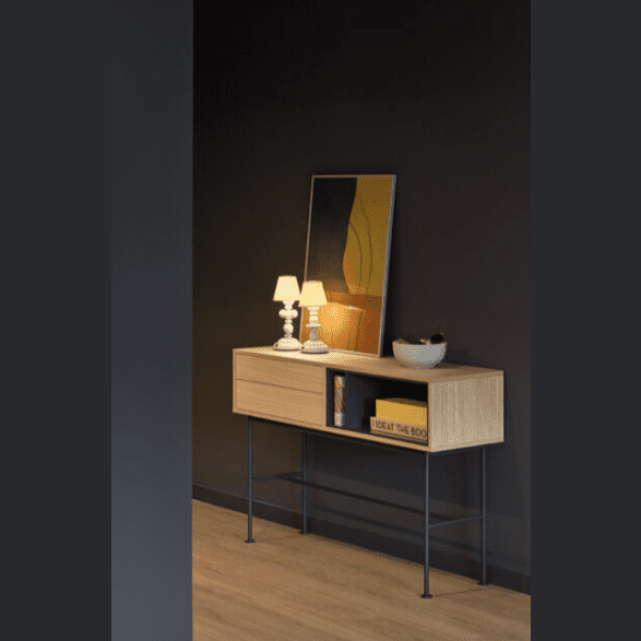 Una mesa auxiliar moderna con acabado de madera similar a la Consola Yoko 2C se coloca contra una pared oscura, adornada con una lámpara encendida, un objeto decorativo y una obra de arte enmarcada en la parte superior. Un libro y un cuenco están cuidadosamente colocados en el estante inferior para un almacenamiento moderno y elegante.