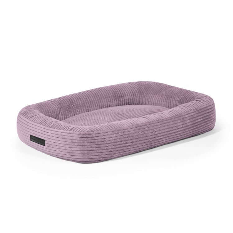 Sobre un fondo blanco se presenta una Cama para perro de terciopelo acanalado 75×60×18 cm - DOG BED 75 WAVES rectangular, de color malva, con bordes elevados y una superficie plana para dormir.