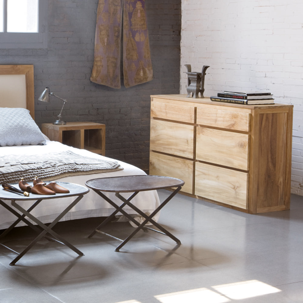 Recámara moderna con cama de madera, mesita de noche a juego, Cómoda con 6 Cajones de Madera Teca Pelikan para organización y estilo, dos taburetes plegables, una lámpara, libros y zapatos sobre una mesa baja contra paredes de ladrillo gris y blanco.
