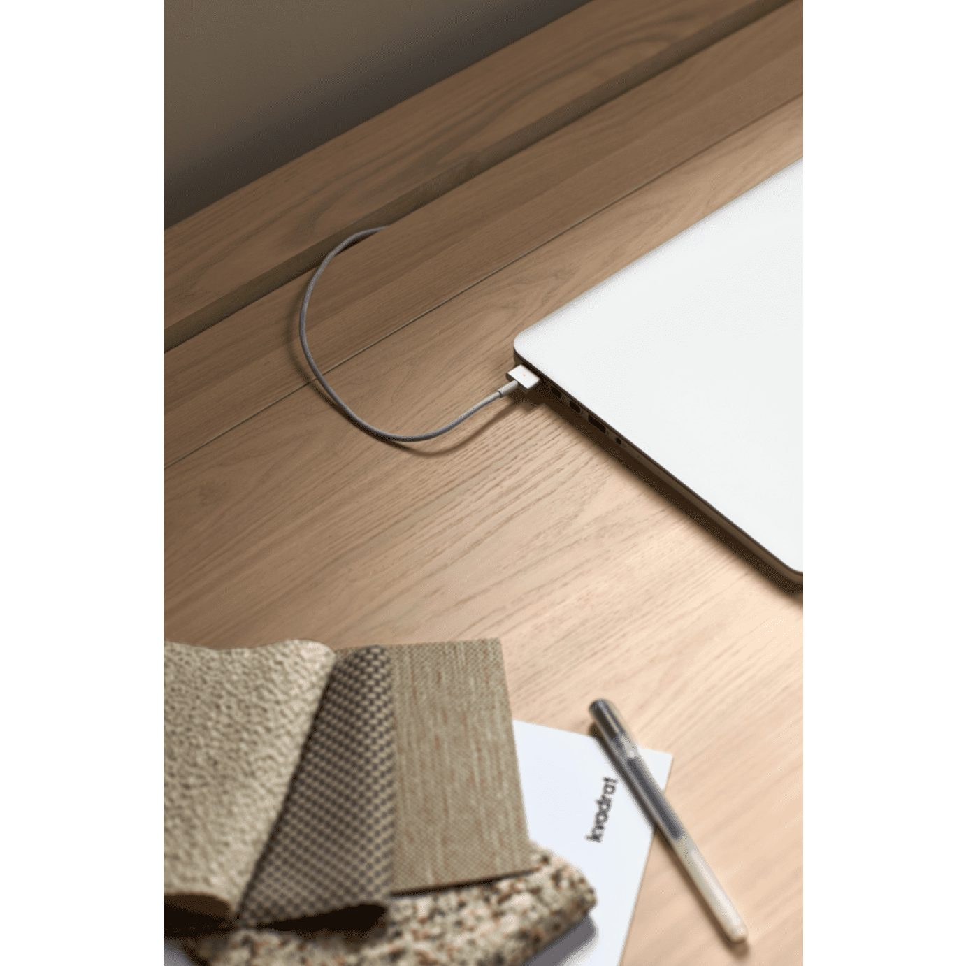 En el Escritorio de Madera Atlas hay una computadora portátil conectada a un cable. Cerca hay muestras de tela, un bolígrafo y una libreta, todo lo cual contribuye a crear un ambiente de diseño moderno con toques de estilo contemporáneo.