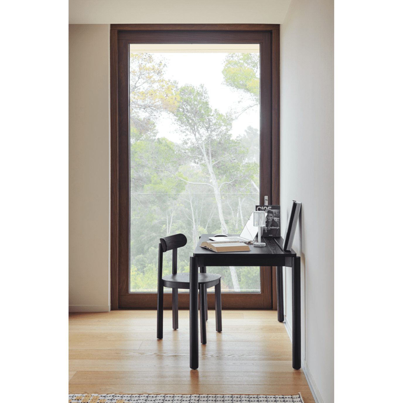 Un rincón de estudio minimalista con un diseño moderno y elegante presenta el Escritorio de Madera Atlas, un pequeño escritorio negro y una silla junto a una gran ventana. Tiene espacio para libros, una cámara, una foto enmarcada y un cuaderno, con árboles visibles afuera.