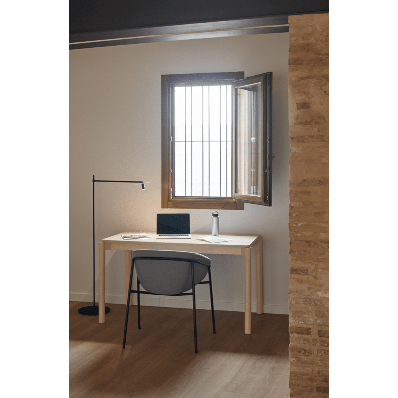 Un espacio de trabajo minimalista exhibe un Escritorio de Madera Atlas, una silla elegante, una lámpara moderna y una computadora portátil junto a una ventana abierta, en una habitación con paredes de piedra y blancas que encarnan el diseño moderno.
