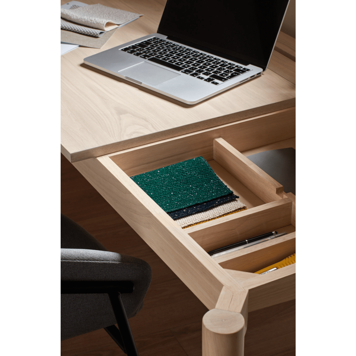 El escritorio de madera Atlas, con un cajón abierto con muestras de tela y bolígrafos, refleja un diseño moderno. Sobre el escritorio hay una computadora portátil parcialmente visible, mientras que una silla cuidadosamente ubicada a su lado rezuma estilo contemporáneo.