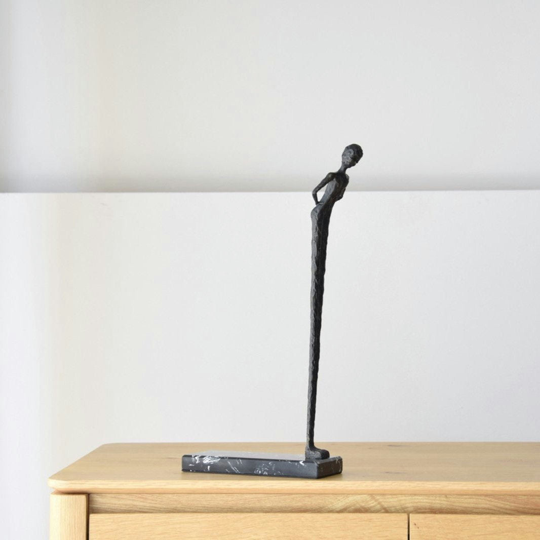 La Escultura Buscando en Metal y Mármol, una esbelta figura abstracta de bronce inclinada hacia adelante, adorna una superficie de madera contra una pared lisa, encarnando una elegancia atemporal que eleva cualquier diseño de interiores.