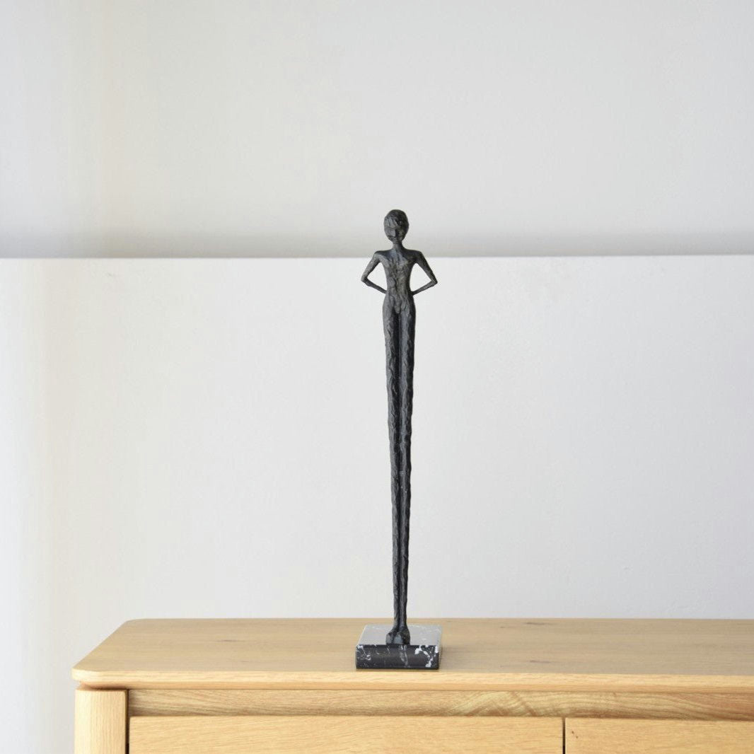 La Escultura Buscando en Metal y Mármol es una escultura negra alta y esbelta de una persona con los brazos en jarras sobre una superficie de madera. Encarnando la elegancia atemporal, es ideal para el diseño de interiores, agregando sofisticación a cualquier espacio.