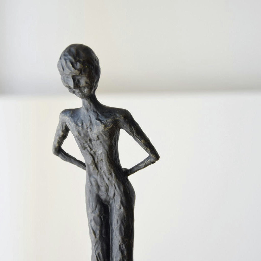 La Escultura Buscando en Metal y Mármol presenta una figura humana esbelta y abstracta con las manos en las caderas sobre un fondo liso, encarnando una elegancia atemporal que eleva cualquier espacio de diseño de interiores.