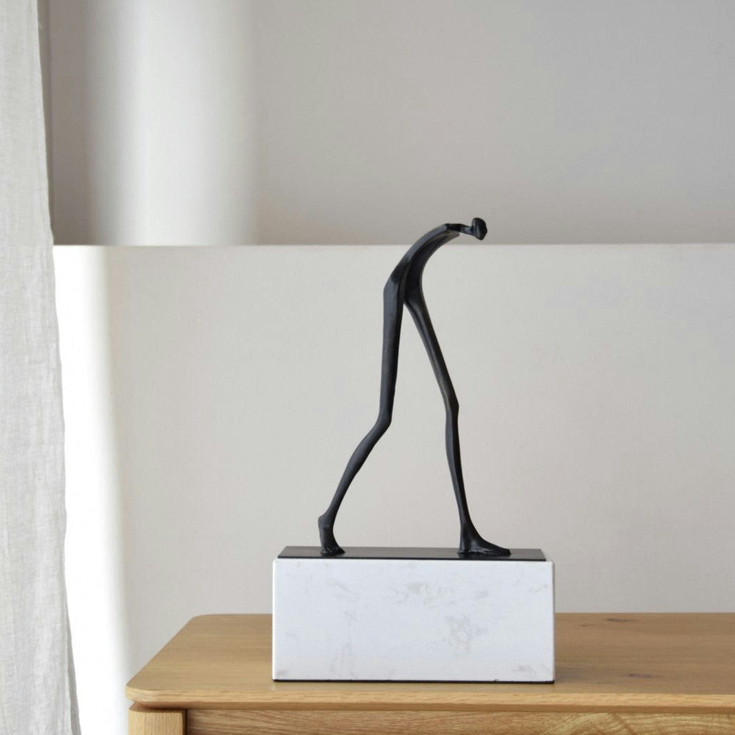 La "Escultura Caminante", una escultura de metal negro de una esbelta figura caminando sobre una elegante base de Mármol Blanco, se asienta elegantemente sobre una superficie de madera, realzando cualquier ambiente de decoración e interiorismo.