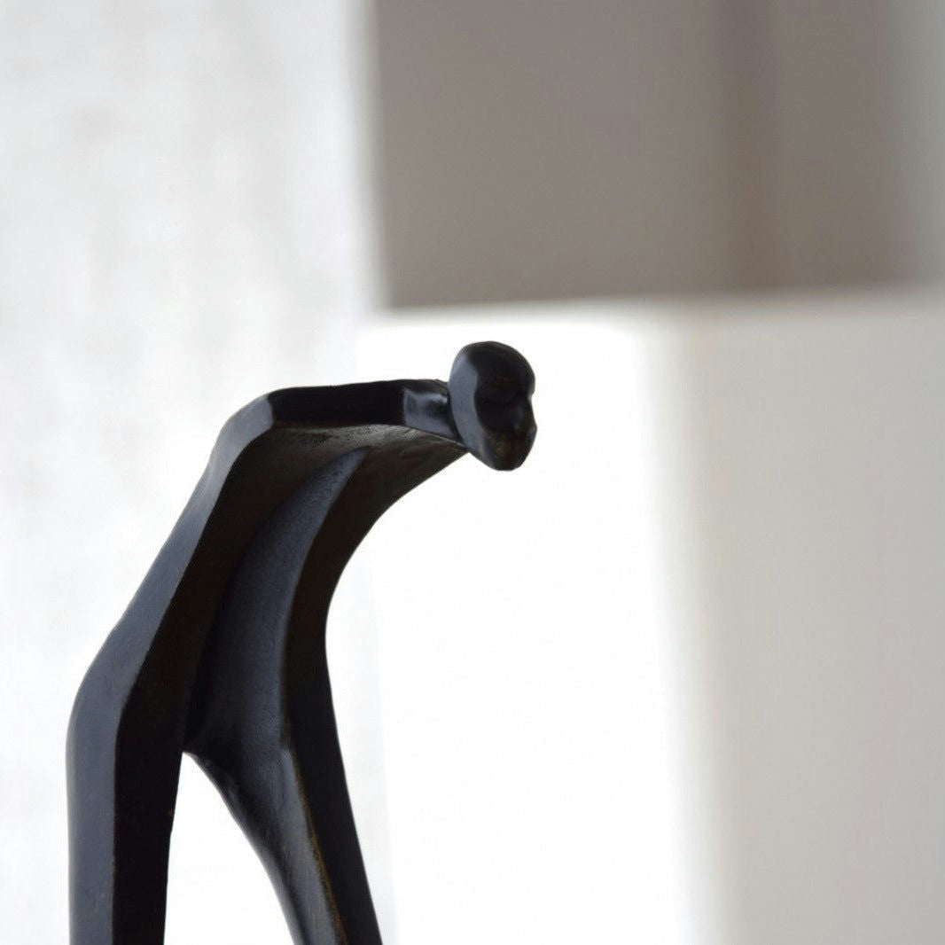 Elegante escultura moderna de color negro de una figura humanoide inclinada hacia delante sobre un fondo neutro, que recuerda la elegancia y sofisticación que se encuentran en Escultura Caminante. Metal y Mármol.
