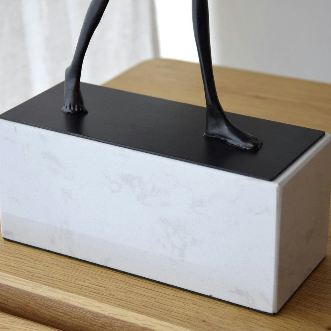 La Escultura Caminante. Metal y Mármol presenta pies de bronce envejecido sobre una base rectangular de mármol blanco, bellamente colocados sobre una superficie de madera.