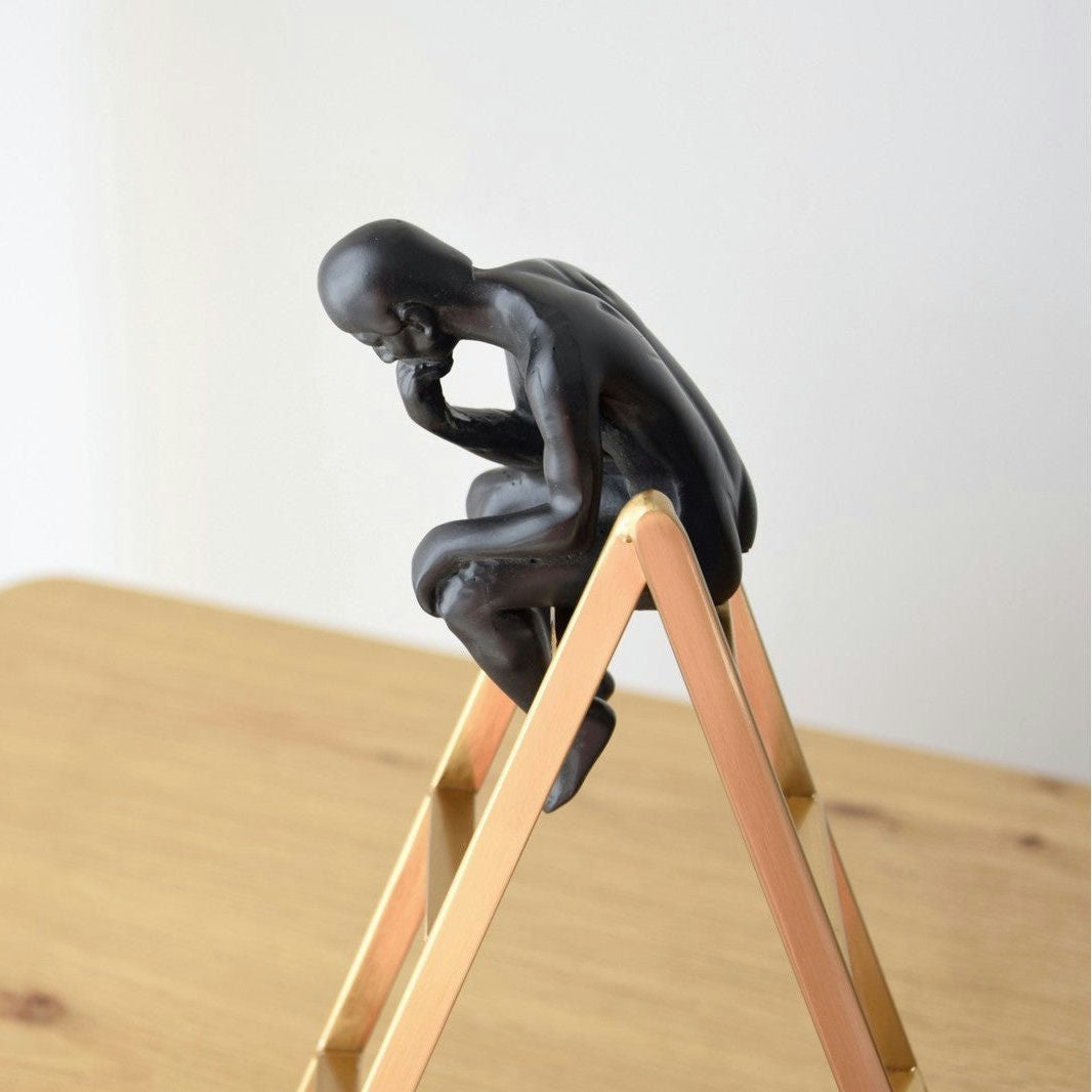 La Escultura de Pensador sobre Escalera es una pequeña escultura de metal negro de una figura contemplativa en una escalera de madera, ideal para agregar sofisticación a su diseño de interiores.