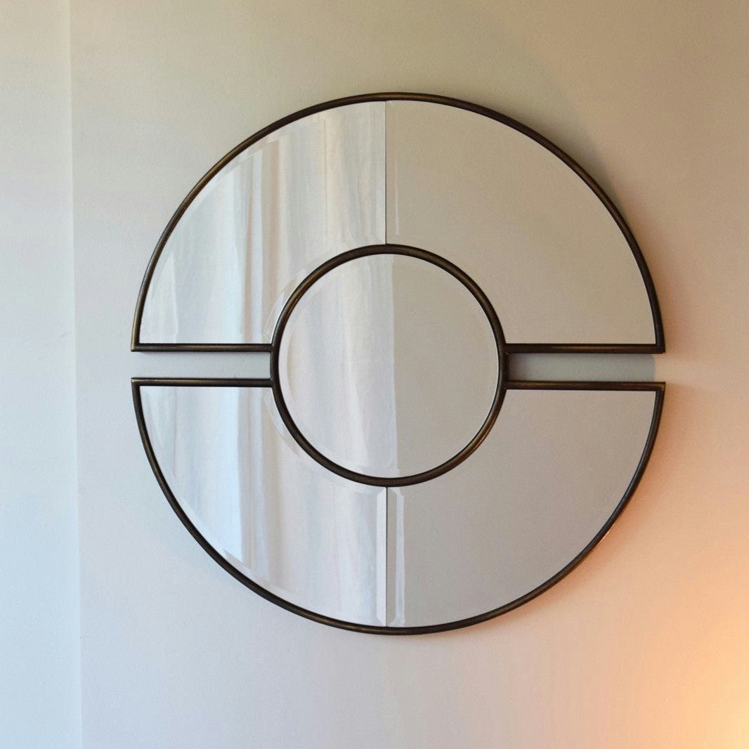 El Espejo Redondo Reflejo Doré presenta un marco metálico y está dividido en cuatro secciones como una Pokeball simplificada, agregando diseño y funcionalidad mientras cuelga elegantemente en una pared de color claro.