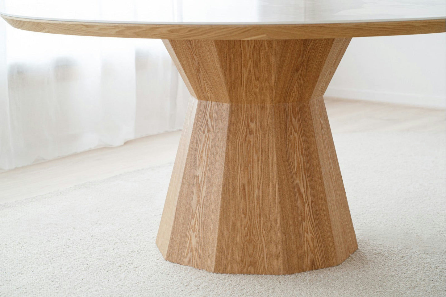 La mesa de comedor redonda en cerámico y fresno de Nat Dread presenta una mesa redonda con una base de madera facetada hecha de cerámica y fresno, cubierta con vidrio. Se encuentra elegantemente sobre una alfombra clara en una habitación luminosa con cortinas blancas transparentes.