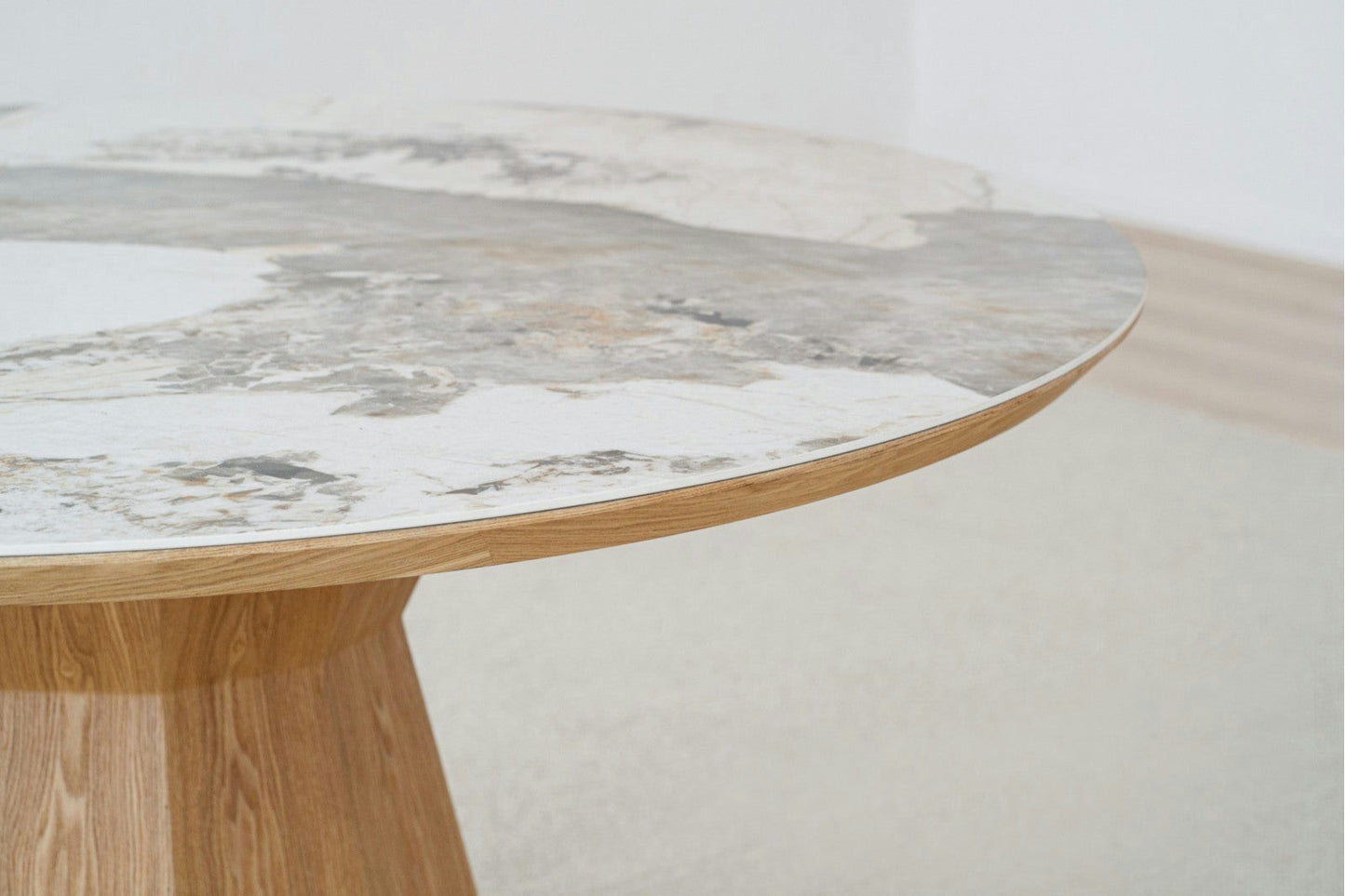 Primer plano de una mesa de comedor redonda Nat Dread con base de madera clara y tapa de cerámica que presenta elegantes patrones en gris y blanco.