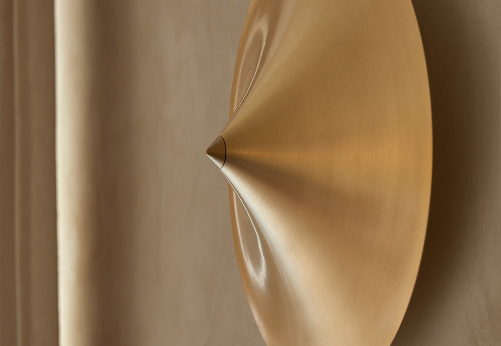 Un primer plano del Aplique de pared Hat, un aplique metálico de forma cónica con un acabado dorado cepillado, colocado sobre un fondo beige, que resalta su iluminación moderna y su estilo de lámpara minimalista.