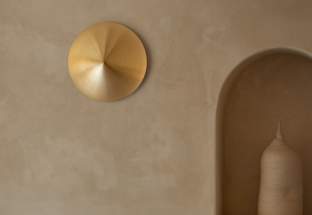 El Aplique de pared Hat, un aplique dorado de forma cónica, está montado en una pared de textura beige junto a una hornacina arqueada con una pieza decorativa alta, aportando iluminación moderna al espacio.