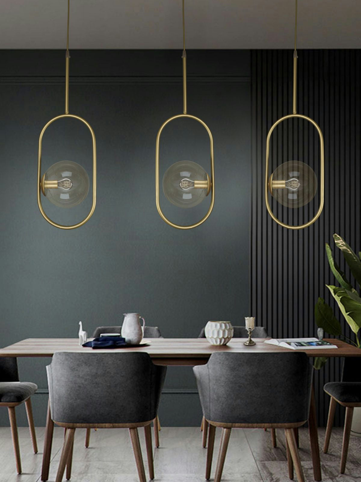 Un comedor moderno que muestra un diseño industrial chic, resaltado por tres Lámpara de Techo Colgante Moderna Dorada 3135-1C-16-3. Una mesa de madera con platos y cuatro sillas grises complementan el espacio, mientras que una planta en maceta agrega elegancia en la esquina contra una pared verde oscuro.