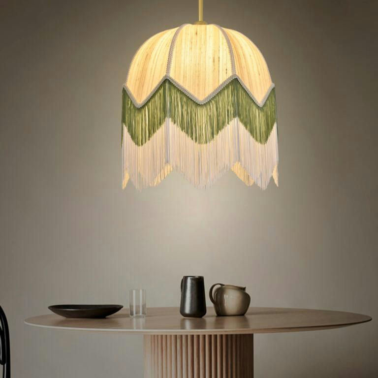 Una mesa redonda de madera con un plato negro, vidrio transparente y dos jarras se encuentra debajo de la Lámpara de Techo Colgante Vintage de Flecos 30148, que presenta una cúpula con flecos en verde y beige para una elegante iluminación difusa.