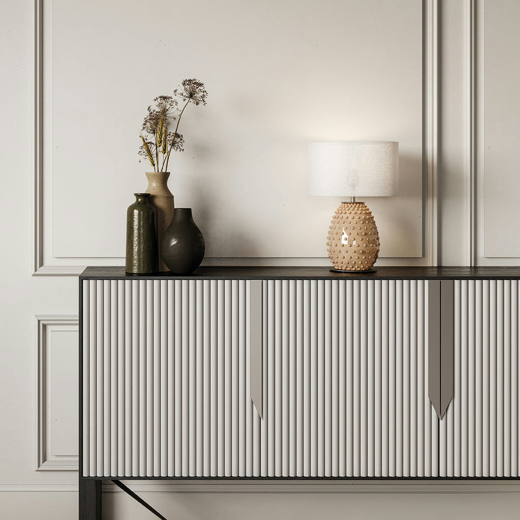 Una consola moderna con listones verticales contra paredes revestidas de paneles blancos encarna el estilo contemporáneo, adornada con jarrones, flores secas y la lámpara de mesa texturizada Cactus NAC135/1.