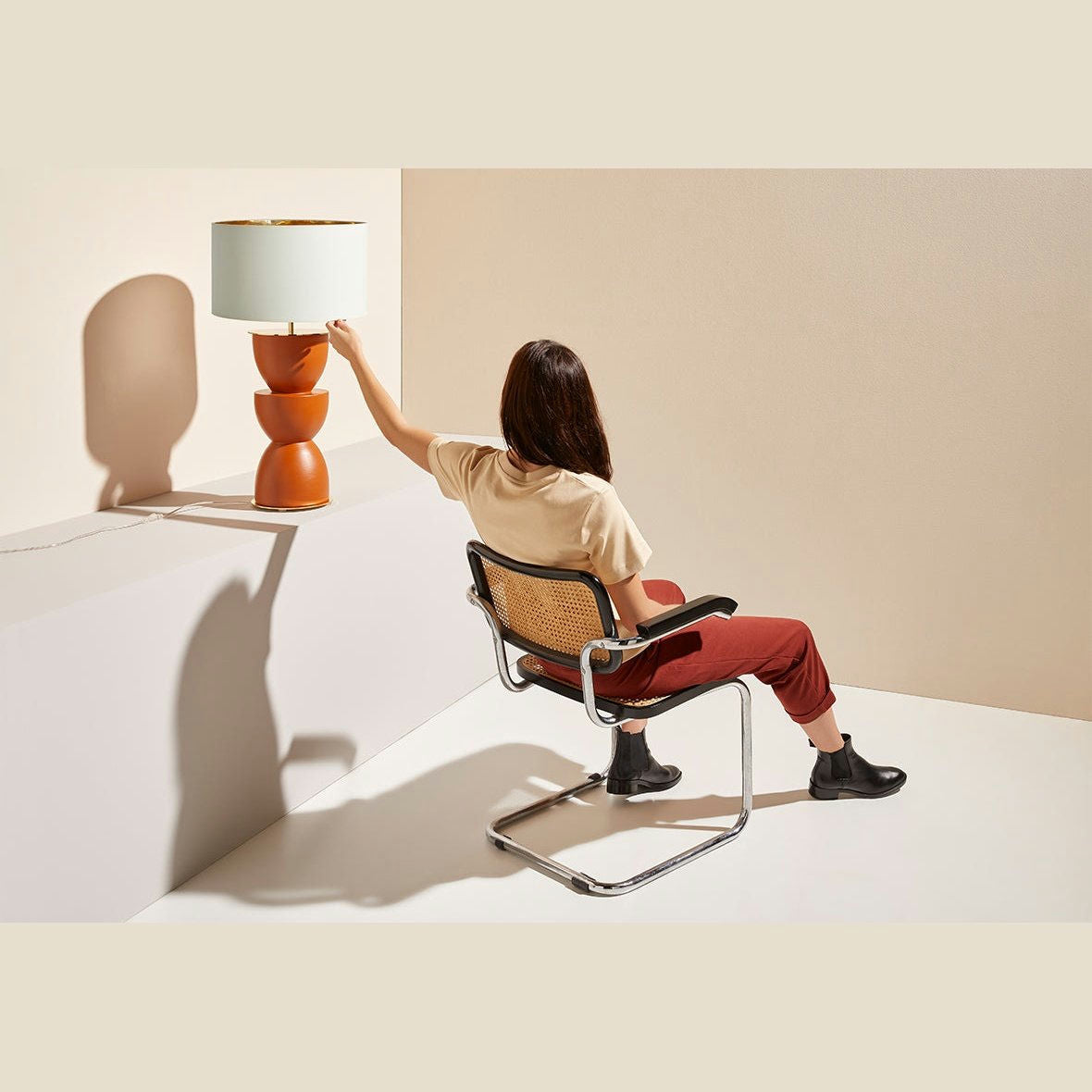 Una persona sentada en una silla de mimbre acomoda una lámpara de mesa Metric sobre una mesa beige. Lleva pantalones rojos y una camiseta beige, que complementan perfectamente la decoración moderna con paredes y suelo en tonos neutros.