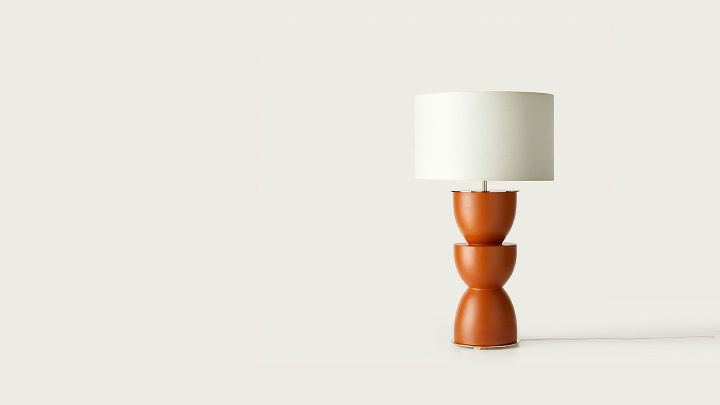 La lámpara de mesa Metric presenta una pantalla cilíndrica blanca y una base geométrica de terracota, que se destaca elegantemente sobre un fondo gris claro, ejemplificando una decoración moderna.