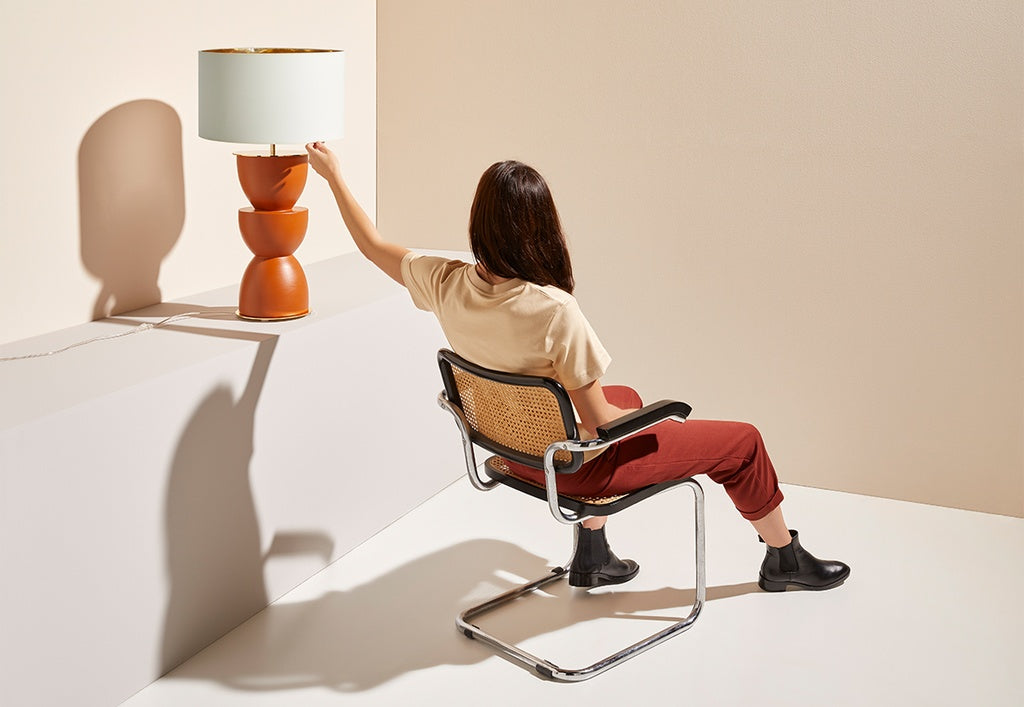 Una persona de cabello largo, sentada en una silla, enciende la Lámpara de mesa en Cerámica y Metal Metric de pantalla blanca y base marrón, sobre una superficie blanca y pared beige, aportando un toque moderno al espacio.