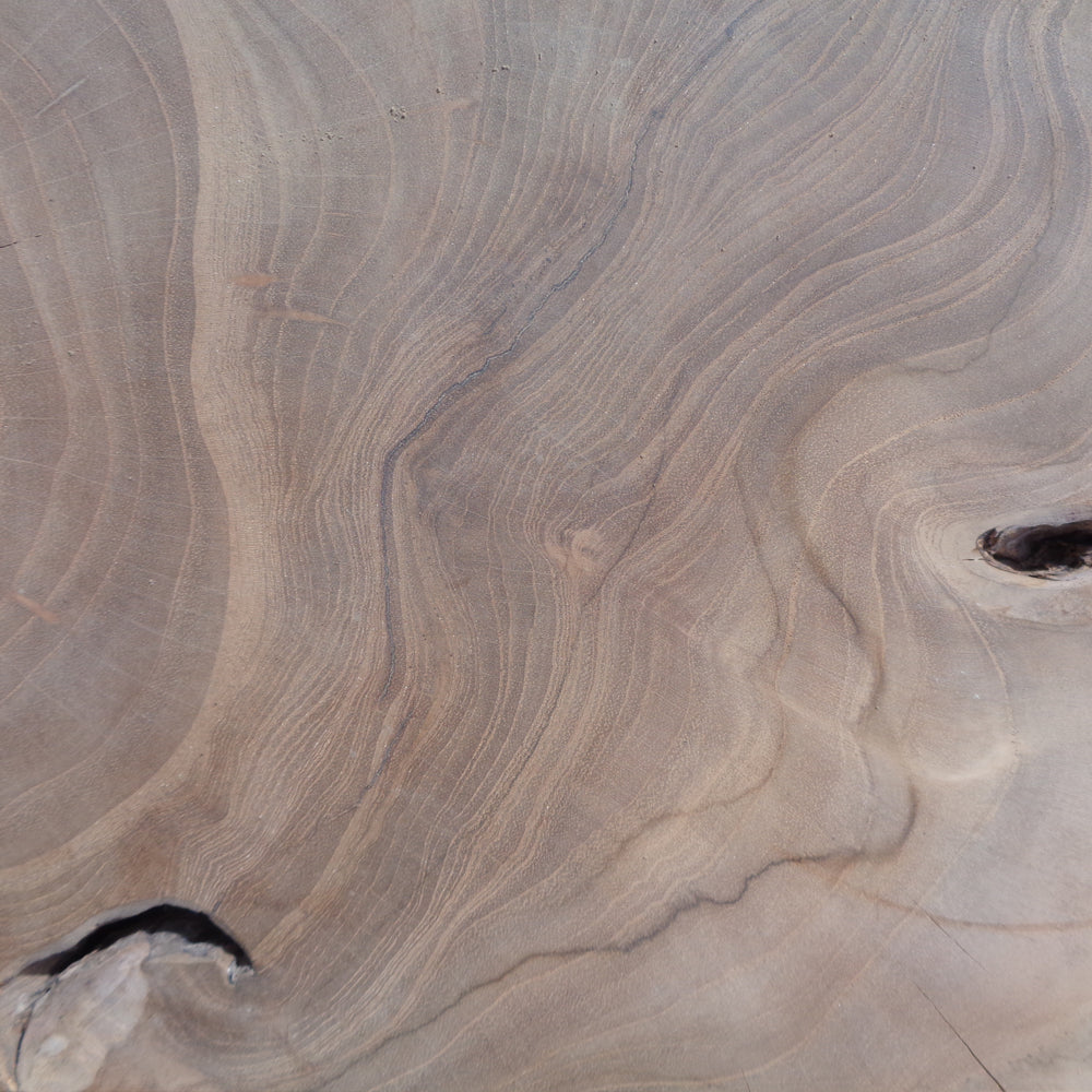 El Puff cuadrado de madera natural Erosion presenta un primer plano de la veta de la madera de teca con patrones claros y oscuros que resaltan su belleza natural. El diseño artesanal incluye pequeños nudos que le aportan carácter a la pieza.