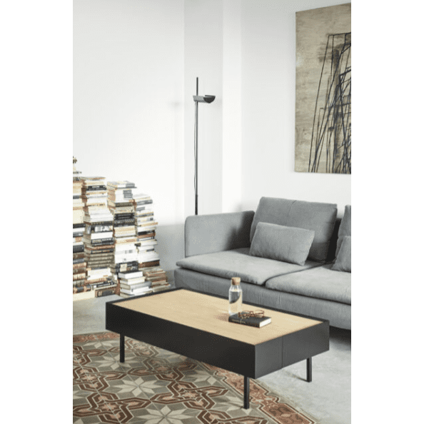 Una sala de estar moderna cuenta con un sofá gris, una pila de libros y una lámpara de pie junto a la Mesa de Centro Arista 2C sobre una alfombra estampada. El arte abstracto cuelga de la pared blanca, mostrando un diseño inteligente.