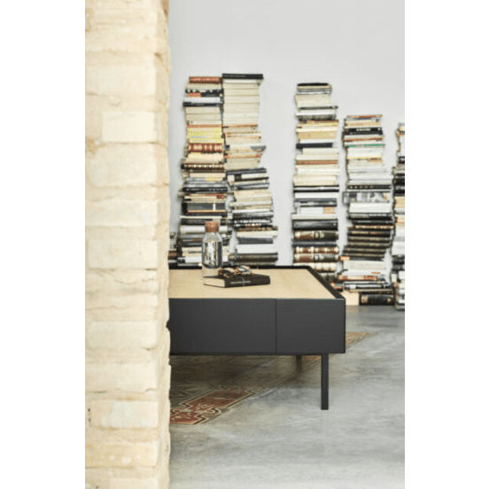 Una habitación minimalista exhibe pilas de libros, la elegante Mesa de Centro Arista 2C con una botella de vidrio y pequeños objetos, y una vista parcial de un pilar de piedra, que encarna el diseño inteligente a través de su elegante organización.