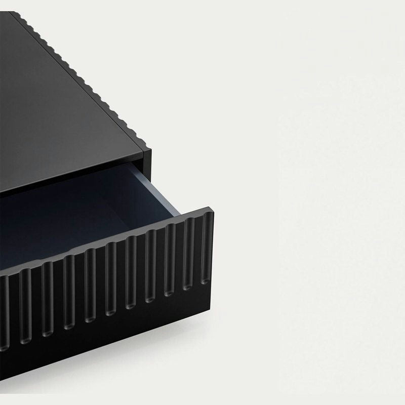 La Mesa de Centro Doric, un cajón negro abierto con textura ranurada que recuerda al diseño neoclásico, proporciona almacenamiento oculto y se extiende desde el lado izquierdo sobre un fondo claro y liso.