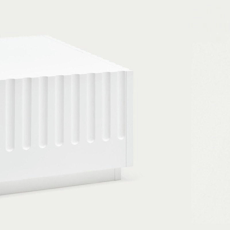 Primer plano de la esquina de la Mesa de Centro Doric, una estructura geométrica blanca con ranuras verticales inspirada en el diseño neoclásico, sobre un fondo claro y liso.