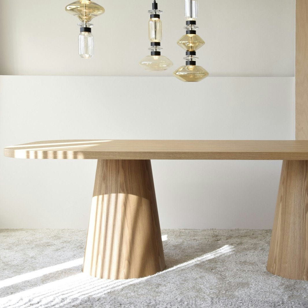 La Mesa de Comedor Fresno Natural Dolmen cuenta con un tablero de madera con patas cónicas sobre una alfombra clara, realzada por luces colgantes. Su chapa de madera natural aporta calidez al diseño minimalista, creando un ambiente elegante y sencillo.