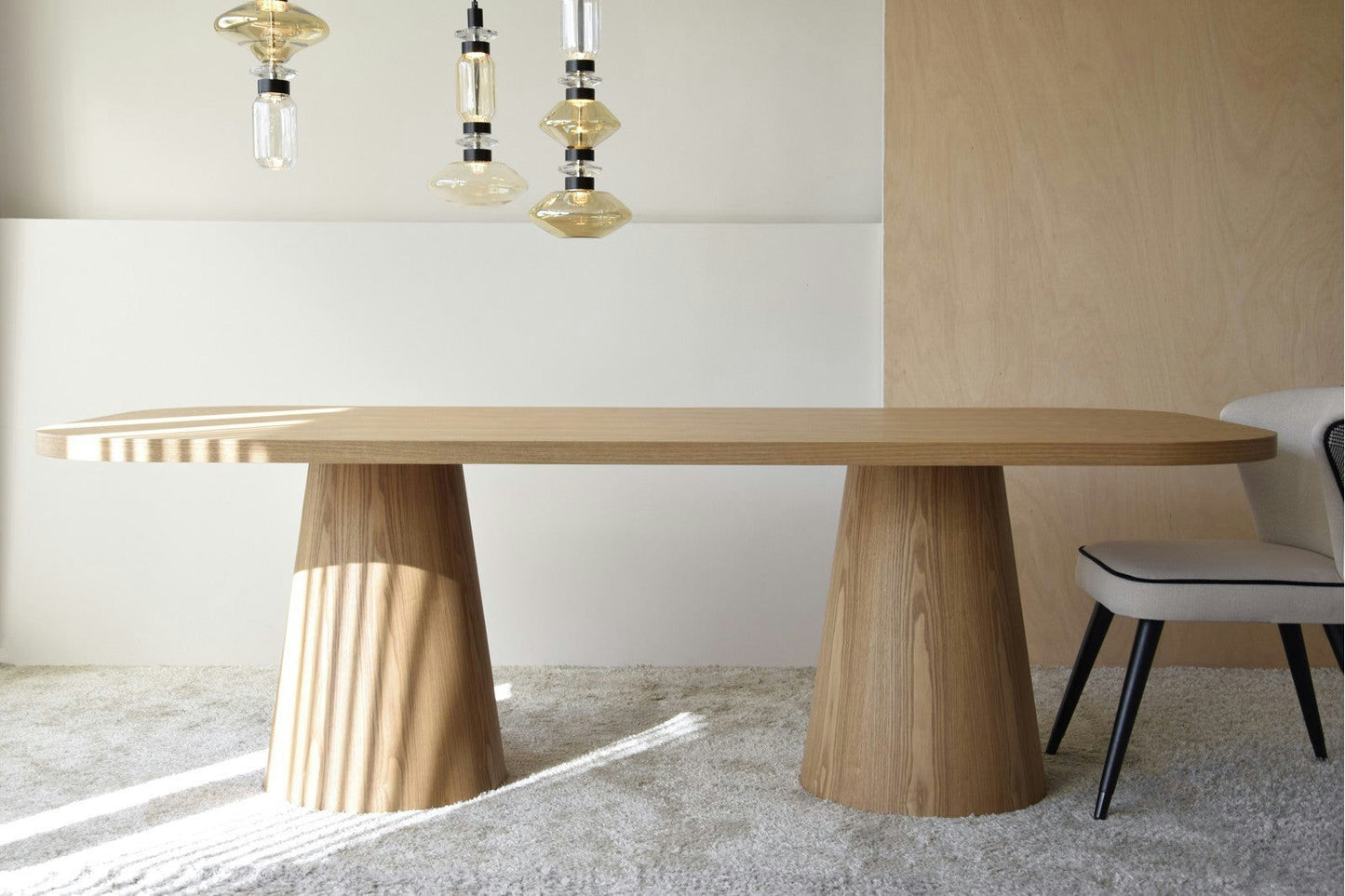 La Mesa de Comedor Fresno Natural Dolmen presenta un diseño de dolmen distintivo con dos patas de pedestal. Perfecta para una habitación minimalista, se asienta sobre un piso alfombrado y está iluminada por tres luces colgantes en el techo. Elaborada con chapa de madera natural, aporta elegancia orgánica al espacio.