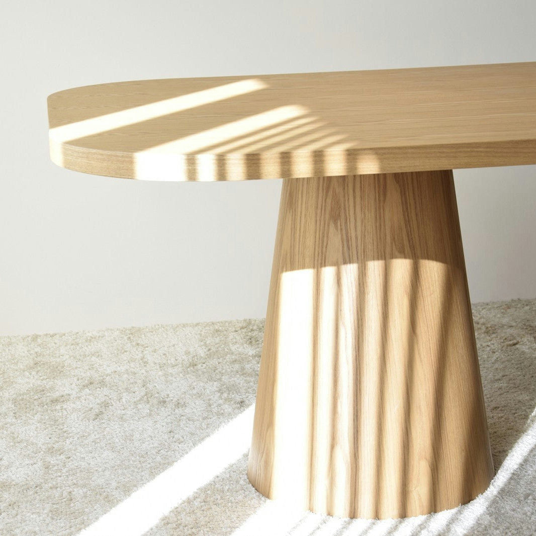 La Mesa de Comedor Fresno Natural Dolmen, una mesa de madera con tapa ovalada y base en forma de cono, está bañada por la luz solar diagonal que proyecta sombras encantadoras sobre su superficie y el suelo.