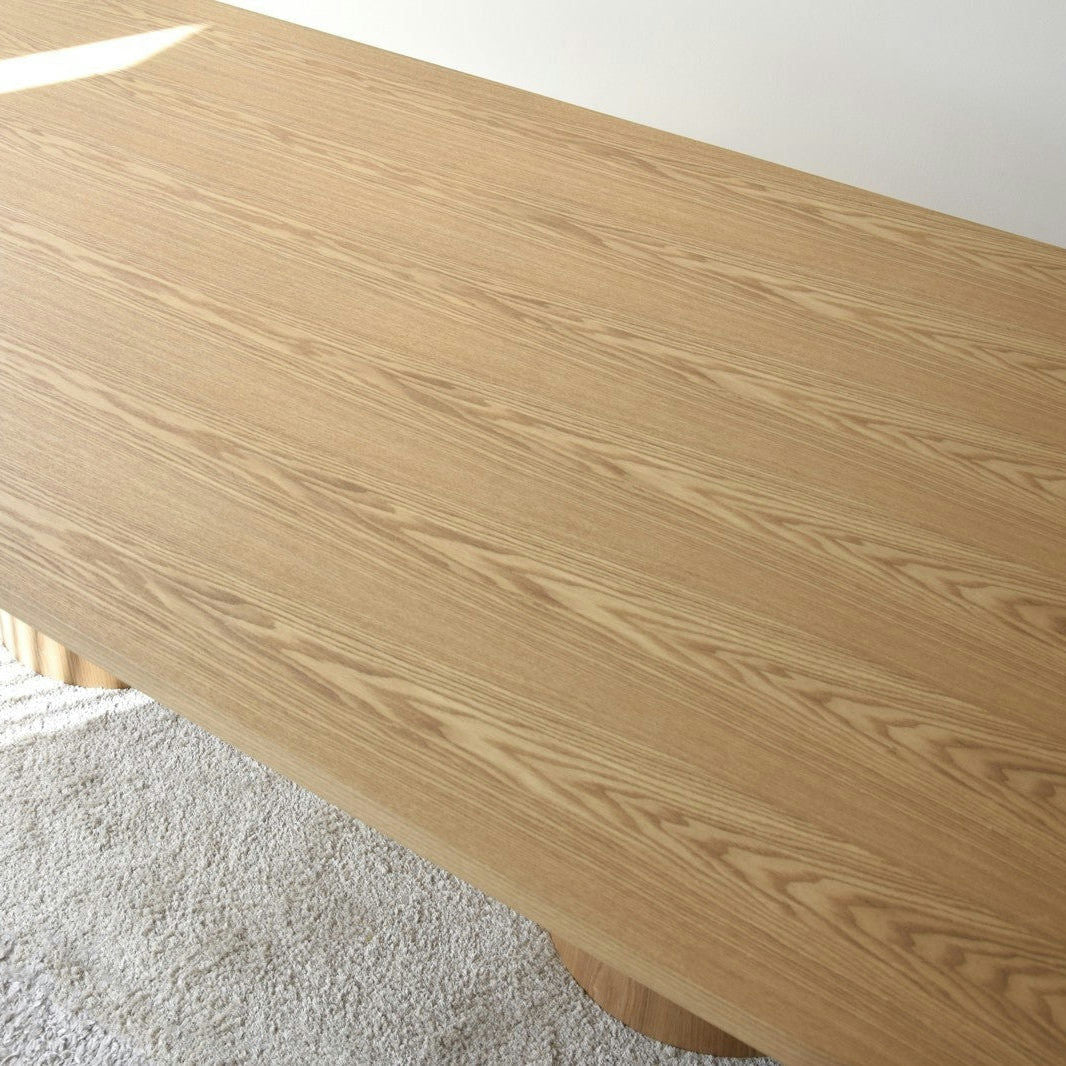 El tablero de madera, en su acabado claro y natural, recuerda a la Mesa de Comedor Fresno Natural Dolmen y reposa elegantemente sobre un suelo alfombrado de color blanco.