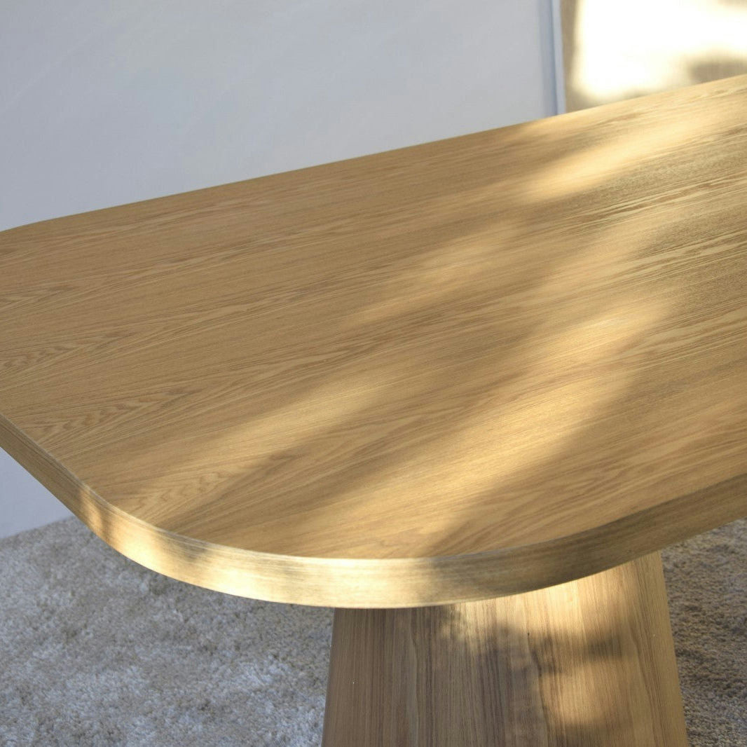 La Mesa de Comedor Fresno Natural Dolmen, con bordes redondeados y chapa de madera natural, irradia elegancia sobre un piso alfombrado bajo una suave iluminación lateral.