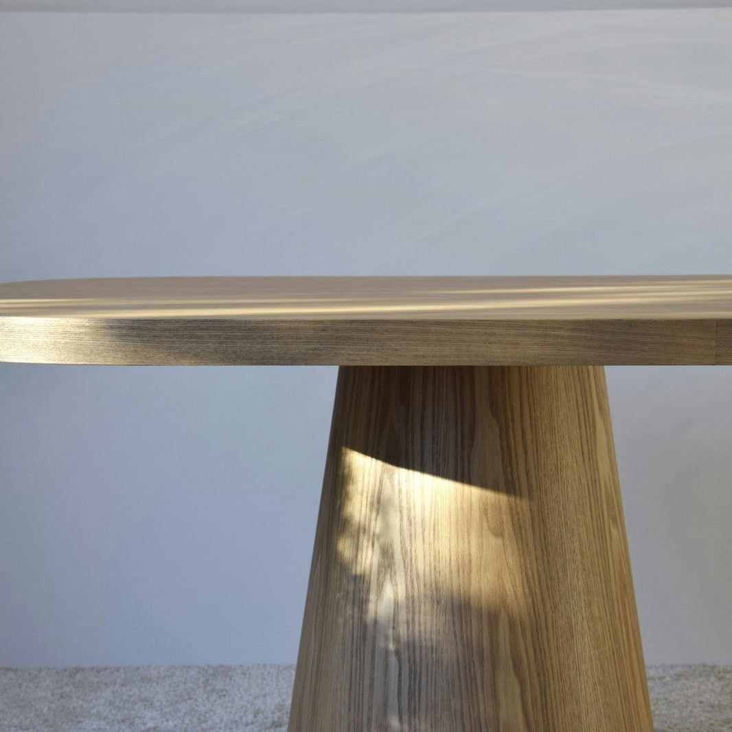 La Mesa de Comedor Fresno Natural Dolmen, con su base redonda cónica y tapa rectangular plana hecha de chapa de madera natural, reposa elegantemente sobre una alfombra de color claro contra una pared lisa, resaltando su diseño único.