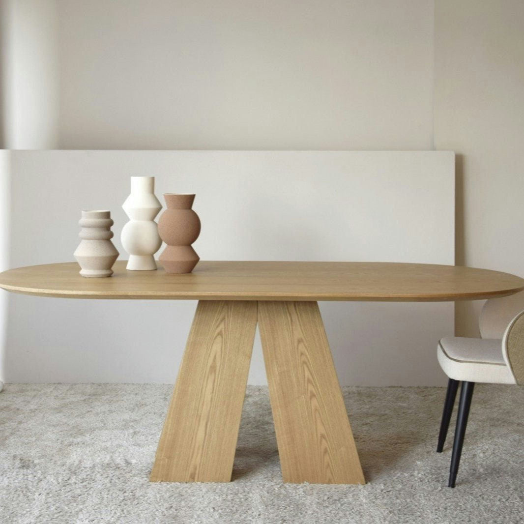 La Mesa de Comedor Fresno Natural Elba presenta un diseño de base único, con tres jarrones esculturales en la parte superior, y está combinada con una silla acolchada que descansa sobre una alfombra clara, que incorpora un toque de diseño sofisticado.