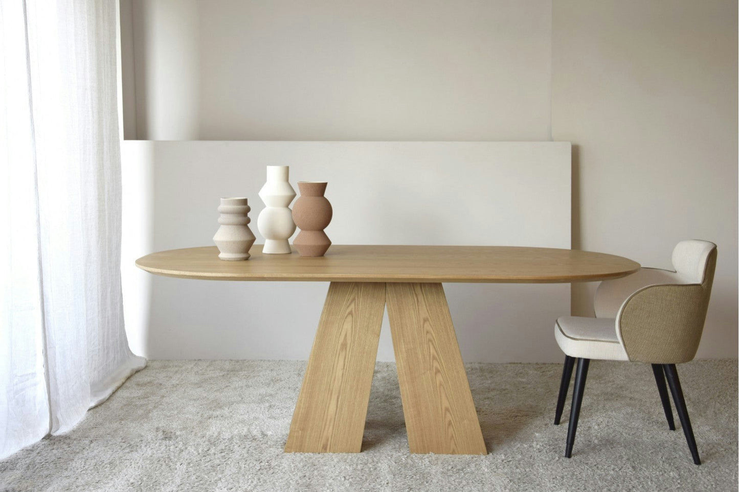 La Mesa de Comedor Fresno Natural Elba con patas geométricas se encuentra sobre una alfombra clara, mostrando un diseño elegante. Tiene tres jarrones de cerámica. A la derecha, una silla con un cojín blanco complementa hermosamente la mesa.