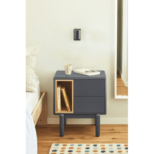 Un dormitorio moderno con una Mesita de Noche Corvo 2C azul marino con libros y una revista, una luz de pared minimalista, una alfombra estampada y destellos de la cama y el espejo, que incorpora un diseño contemporáneo con almacenamiento eficiente.