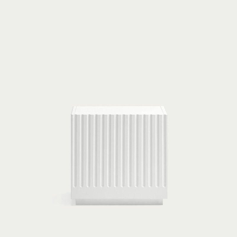 La Mesita de Noche Doric 1C, un pequeño cubo blanco con crestas verticales como columnas dóricas, se destaca elegantemente sobre un fondo gris claro liso, agregando un toque de diseño contemporáneo.