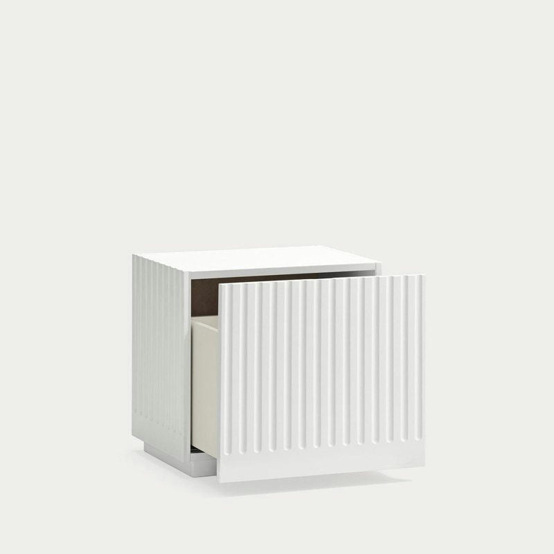 La mesita de noche Doric 1C es una mesita de noche cuadrada de color blanco con una textura de rayas verticales inspirada en las columnas dóricas. Tiene un cajón parcialmente abierto y reposa sobre un fondo claro y liso.