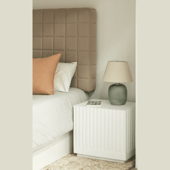 El dormitorio cuenta con una cabecera tapizada en beige, una cama blanca con almohadas en beige y blanco y la elegante Mesita de Noche Doric 1C. Una lámpara con textura verde agrega estilo, mientras que un control remoto se encuentra cerca, combinando a la perfección la elegancia con el diseño contemporáneo.