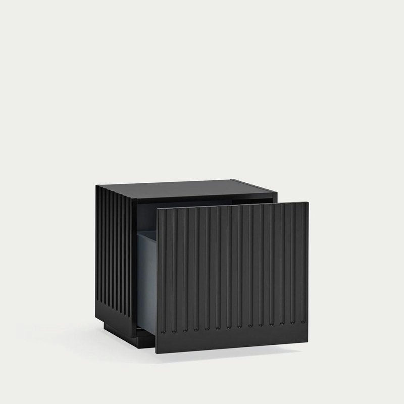 La Mesita de Noche Doric 1C es un mueble negro en forma de cubo con ranuras verticales que recuerdan a columnas dóricas y que ofrece un diseño contemporáneo. Su gran cajón está parcialmente abierto, lo que añade intriga al fondo blanco liso.