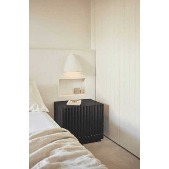 Un dormitorio minimalista incluye una elegante mesita de noche Doric 1C junto a la cama y una lámpara de pared. Un libro y una taza descansan en la alcoba mientras que la ropa de cama y las paredes en tonos crema realzan el diseño contemporáneo para crear una atmósfera serena.