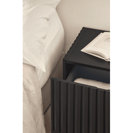 Junto a la cama se encuentra una elegante mesita de noche Doric 1C en color negro, que encarna el diseño contemporáneo. Sobre ella hay un libro abierto y en el cajón hay una toalla cuidadosamente doblada.