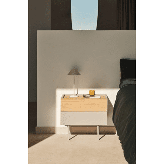 La mesita de noche Otto 2C, una mesa de noche de madera, sostiene una lámpara, velas y libros contra una pared blanca. La luz que entra por una ventana alta proyecta sombras en el piso, lo que resalta su diseño moderno y brinda almacenamiento compacto para sus elementos esenciales.